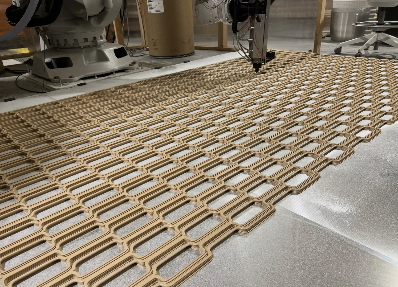 A robotic arm printing a large repeating lattice of interlocking rounded openings in tan filament across a metal build plate